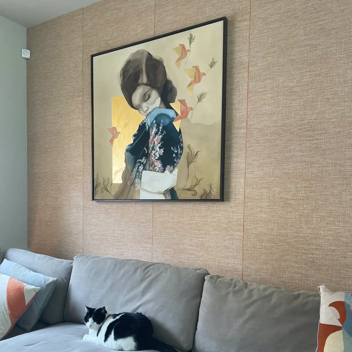 woonkamer muur met licht oranje behang van Arte en een grote kunstwerk met daarvoor een grijze bank met een zwart witte poes.