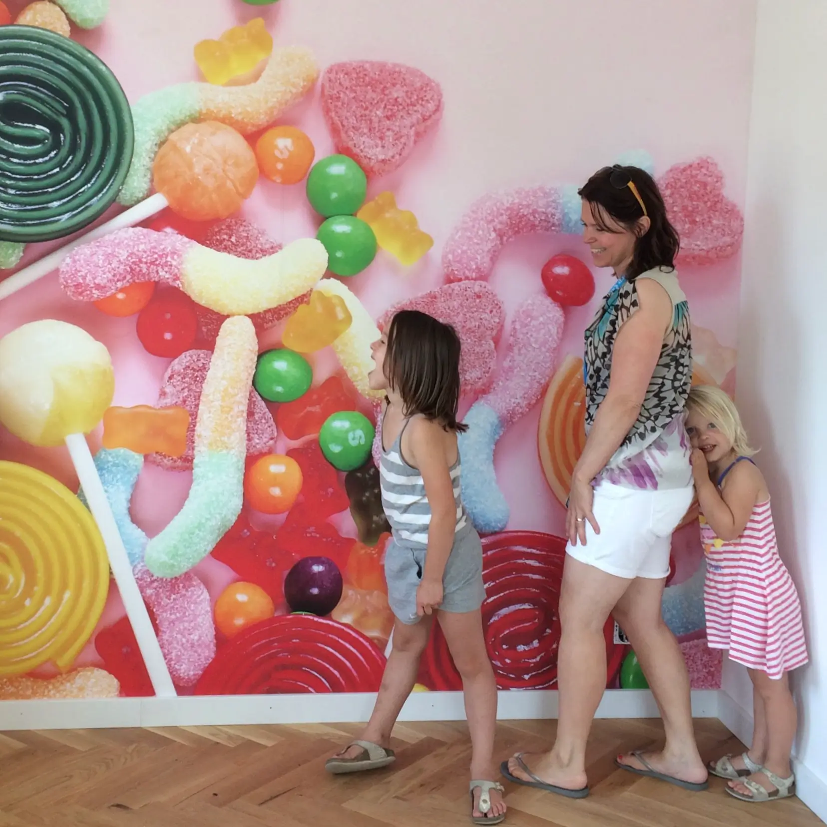 Muur met fotobehang waar grote snoepjes op staan afgebeeld. Voor de muur een blije moeder en twee kinderen.