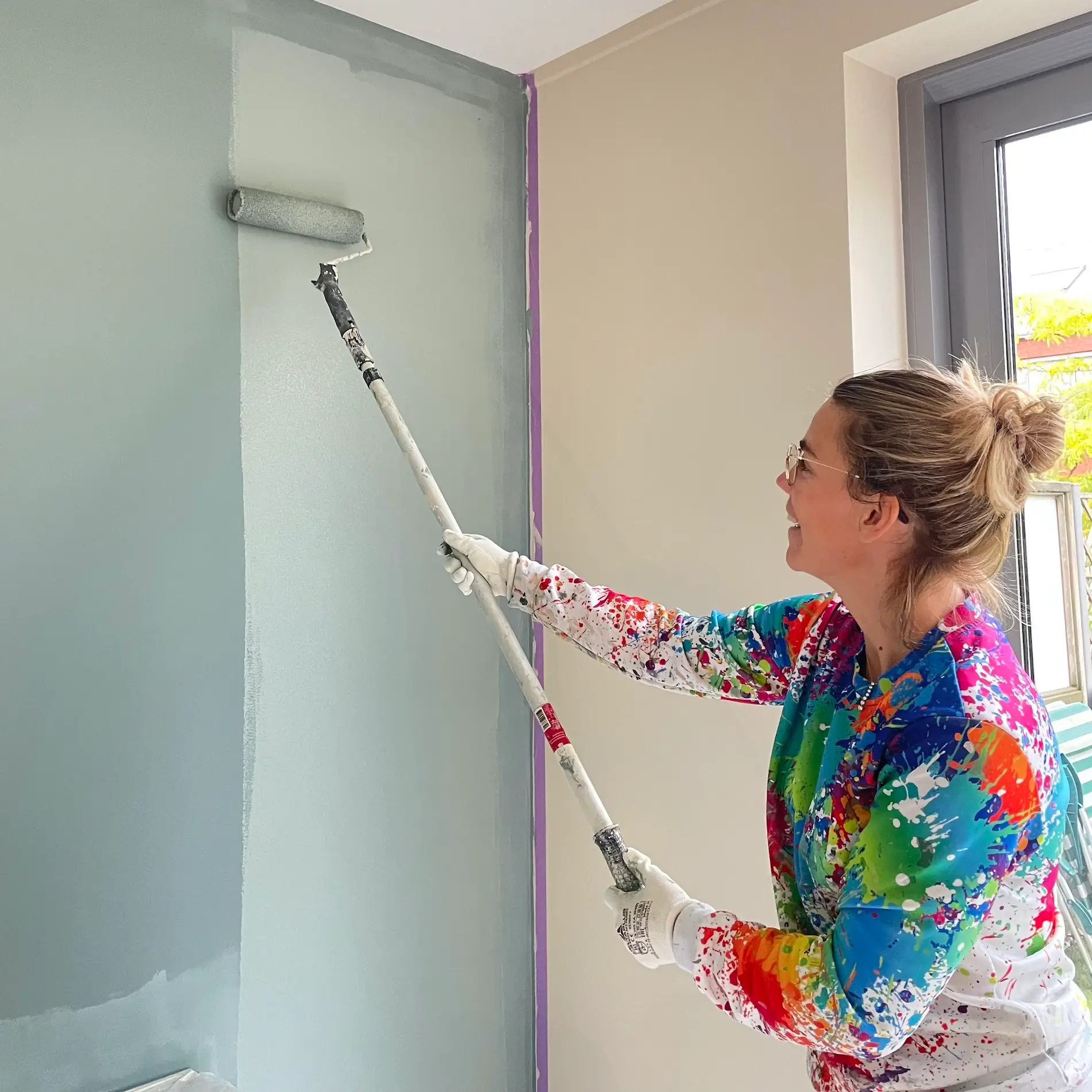 Vrouwelijke schilder verft muur met een roller op een stok. In de hoek zie je tape op de muur zitten.