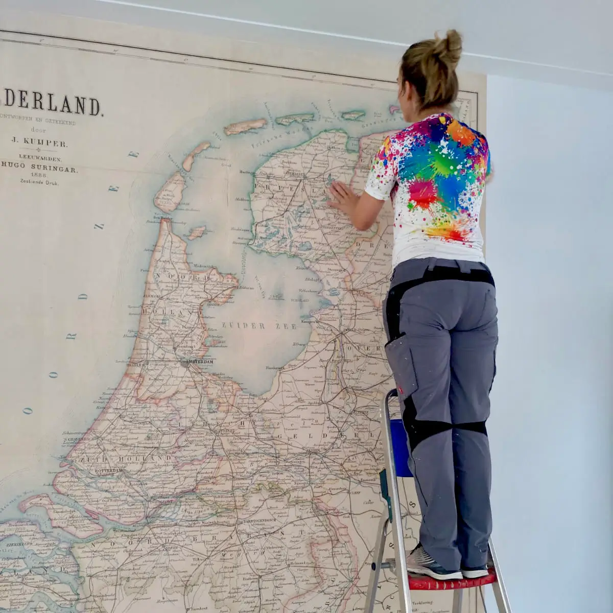 Vrouwelijke behanger die op een trap staat en een ouderwetse landkaart van Nederland aan het behangen is.